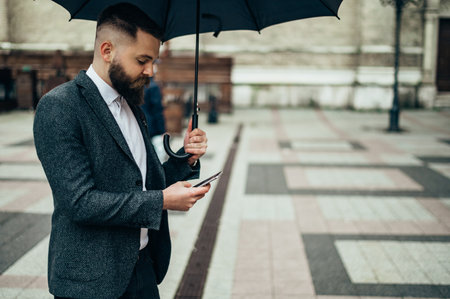 Young handsome businessman using a smartphone and holding a black umbrella while outside on a rainy dayの写真素材