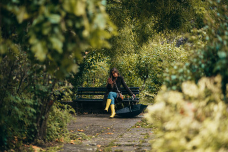 Beautiful young woman using smartphone while sitting on a bench in the park wearing yellow rubber bootsの写真素材