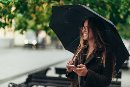 Beautiful young woman using smartphone and holding an umbrella while walking in the city on a rainy dayの写真素材