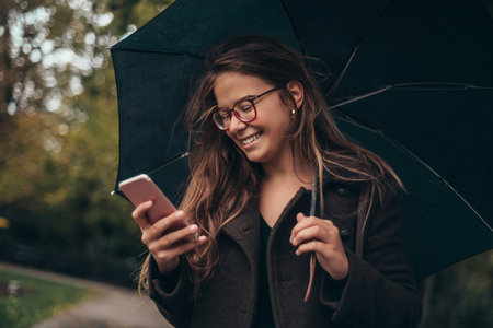 Beautiful young woman using smartphone and holding an umbrella while walking in the city on a rainy dayの写真素材