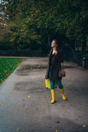 Beautiful young woman holding an umbrella and wearing yellow boots and having a yellow suitcase in the cityの写真素材