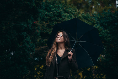 Beautiful young woman enjoying the sound of rain while holding a black umbrella and walking in the parkの写真素材