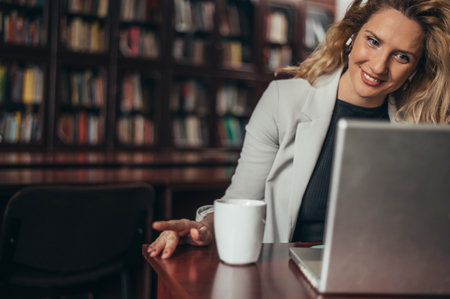 Beautiful young businesswoman using a laptop in her office while drinking coffee and smilingの写真素材