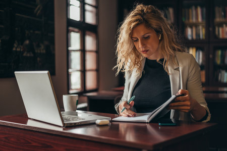 Young beautiful businesswoman using a laptop and a business planner while drinking coffee and working in an officeの写真素材