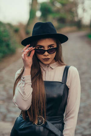 Young fashionable woman posing outside while wearing dress and a hat a holding sunglasses