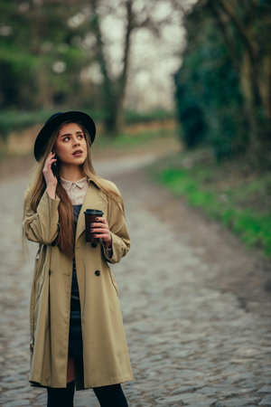 Young fashionable beautiful woman walking on the city streets while using a smartphone and drinking...