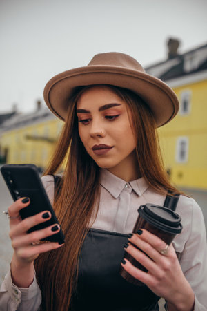 Young beautiful fashionable woman using a smartphone and drinking coffee takeaway while walking...