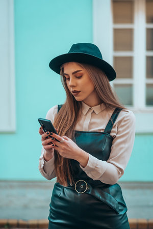 Young fashionable beautiful woman using a smartphone and wearing a hat with the blue wall in the...
