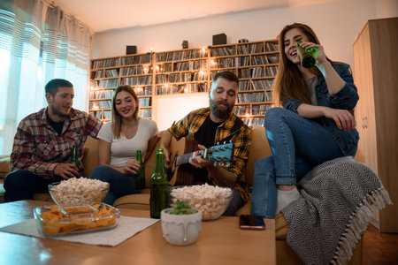 A group of friends on a house party playing guitar and drinking beer while having fun and eating nachos and popcornの写真素材