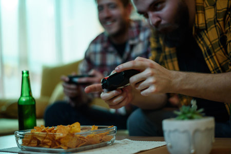 Two best friends playing video games while relaxing at home on the couch and drinking beer and...