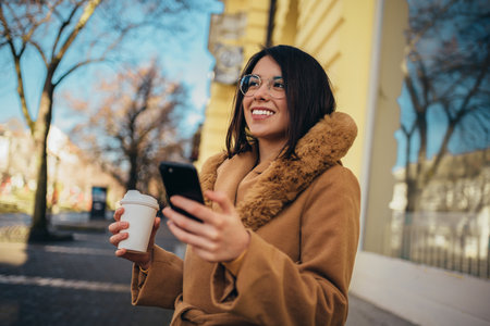 Young hispanic woman using a smartphone and drinking coffee takeaway while walking in the cityの写真素材