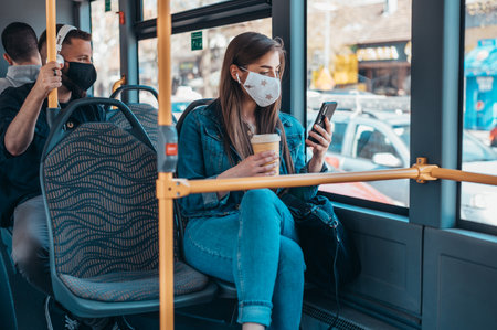 Young woman wearing protective mask riding a bus while using a smartphone and holding a coffee takeaway during corona virus pandemicの写真素材