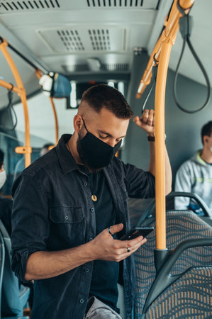 Young man wearing a protective mask while riding the bus and using a smartphone while keeping the distance from other passengers due to corona virus pandemicの写真素材