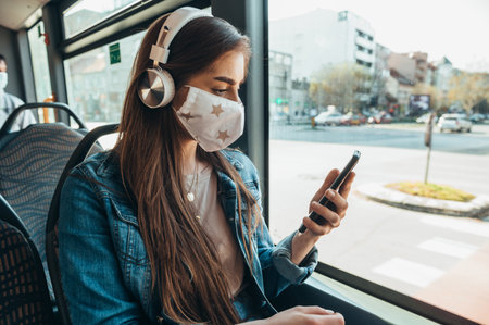 Young beautiful woman wearing protective mask and using a smartphone while riding a bus with a headphones on her headの写真素材