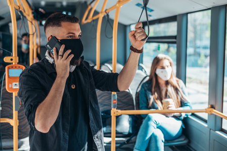 Man wearing protective mask and using a smartphone while riding a bus and standing on a distance from other passengers due to covid19 pandemicの写真素材