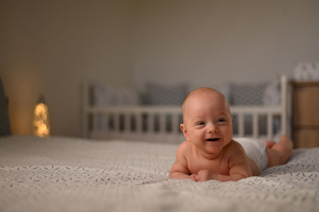Adorable little baby boy lying on his belly on the bed in his room while laughing and wearing only...