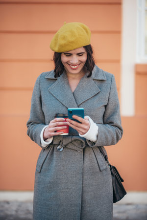 Beautiful young woman using smartphone and wearing yellow beret and a gray coat walking down the city streetsの写真素材