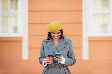 Portrait of an attractive young woman in gray coat wearing a yellow beret and using a smartphone while standing against the orange wallの写真素材