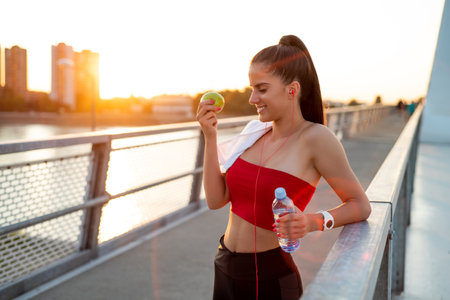 hot of a sporty young woman resting on a bridge after workout eating an apple and drinking waterの写真素材