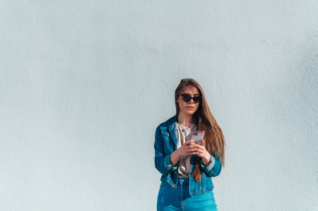 Young beautiful woman using a smartphone while standing in front of a white wall and being lit by the sunの写真素材