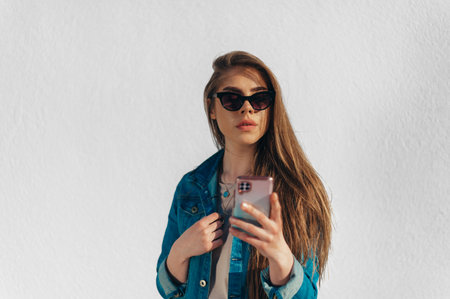 Young beautiful woman using a smartphone and wearing jeans and sunglasses with a white wall in the backgoundの写真素材