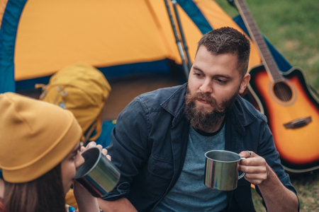 Young couple of campers drinking coffee from metal cups while sitting near the tent in their camp in the nature and having funの写真素材