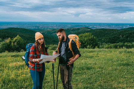 A young couple of hikers using trekking poles and map while wearing backpacks with camping gear and...