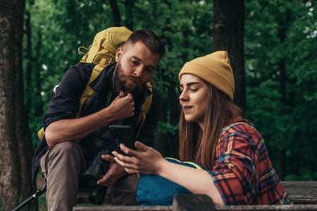 A couple of hikers sitting on a bench in the forest and making a break while using a smartphone for orientation and finding the next pathの写真素材