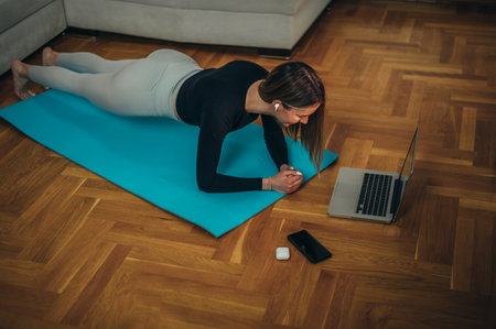 Young attractive woman in a plank position training at home while using a laptop