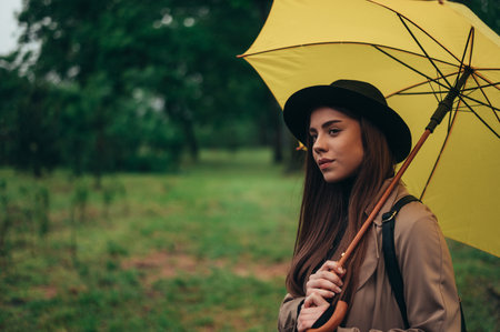 Beautiful young woman holding yellow umbrella while walking in the park on a rainy dayの写真素材