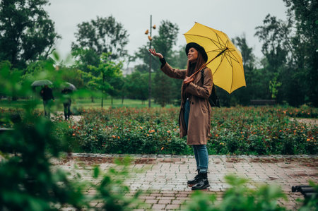 Young beautiful woman using smartphone and holding yellow umbrella while walking in the park during...