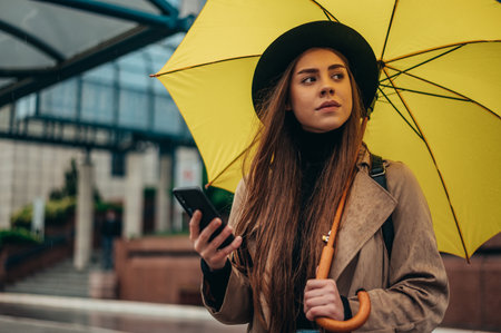 Beautiful young woman using smartphone and holding a yellow umbrella while in the city during rainの写真素材