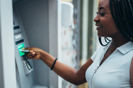 Cheerful african american woman using credit card and withdrawing cash at the ATMの写真素材