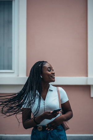 Beautiful african american woman using a smartphone and hair flipping her braids while out in the city by dayの写真素材