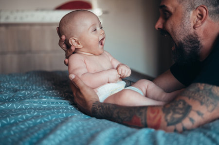 Tattooed bearded father playing with his son in diapers while at home on the bed