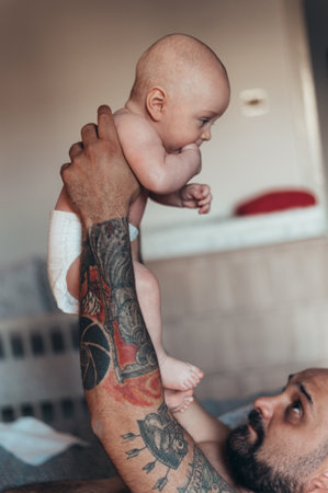 Tattooed bearded father playing with his son in diapers while at home on the bedの写真素材