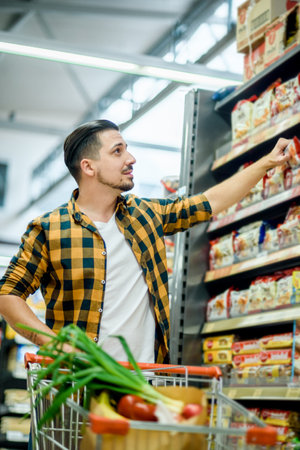 Young handsome man in a supermarket choosing products and holding shopping cart while grocery shoppingYoung handsome man in a supermarket choosing products and holding shopping cart while grocery shoppingの写真素材