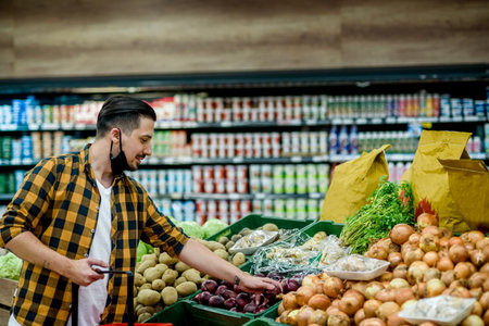 Young handsome man in a supermarket wearing protective mask and choosing products and holding shopping cart while grocery shoppingの写真素材