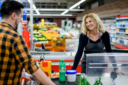Beautiful young cashier is smiling while working at the supermarket and wearing an apronの写真素材