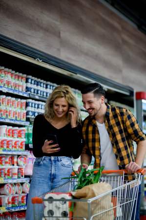 Young happy couple using smartphone in supermarket with shopping cart choosing products while grocery shoppingの写真素材