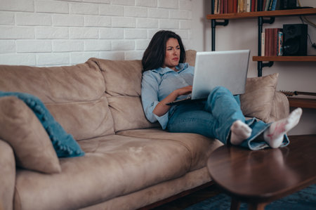 Young businesswoman working on a laptop while relaxing on the couch and working from home with legs raised on a tableの写真素材
