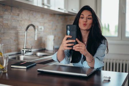 Young businesswoman taking selfie with her smartphone while sitting in her kitchen at home and workingの写真素材