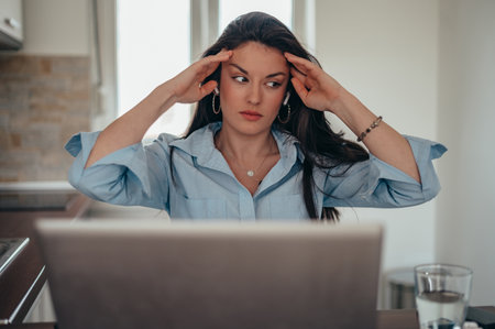 Young tired businesswoman using a laptop and airpods while working from home on the kitchen counter...