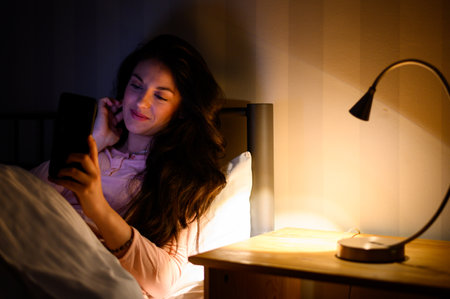 Young beautiful woman lying in her bed during night and using a smartphone while being illuminated...