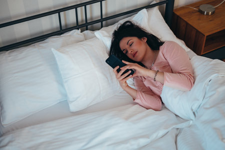 Young beautiful woman waking up in her bed at home while holding a smartphone in her handの写真素材