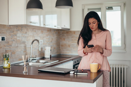 Beautiful young woman wearing a pajama and using a smartphone while working from home and sitting in the kitchenの写真素材