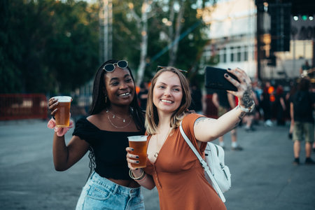 Two beautiful friends taking selfie with a smartphone while drinking beer and having fun at music festivalの写真素材