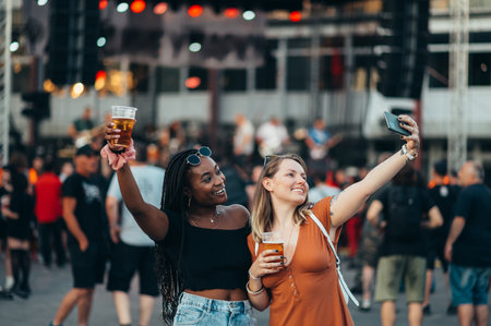 Two handsome friends taking selfie with a smartphone and drinking beer while having fun at music festivalの写真素材