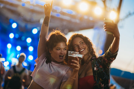 Two beautiful friends taking selfie with a samrtphone while drinking beer and having fun on a music festivalの写真素材