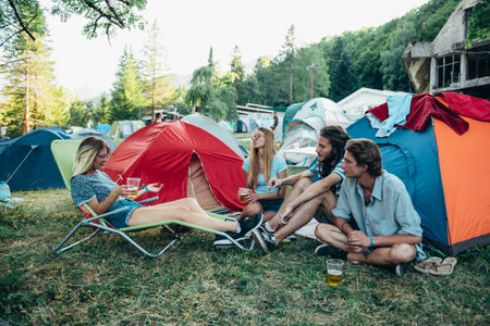 Friends having fun on the campsite at a music festival. Hipsters having fun in their camp near the tents. Young people drinking beer and having a conversation during the day on a summer music festivalの写真素材